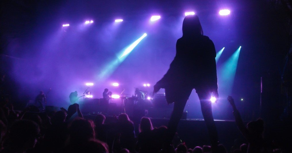 Blue and purple lights shining down onto stage. Shadowed woman in the right foreground. Blurred outline of people in the crowd