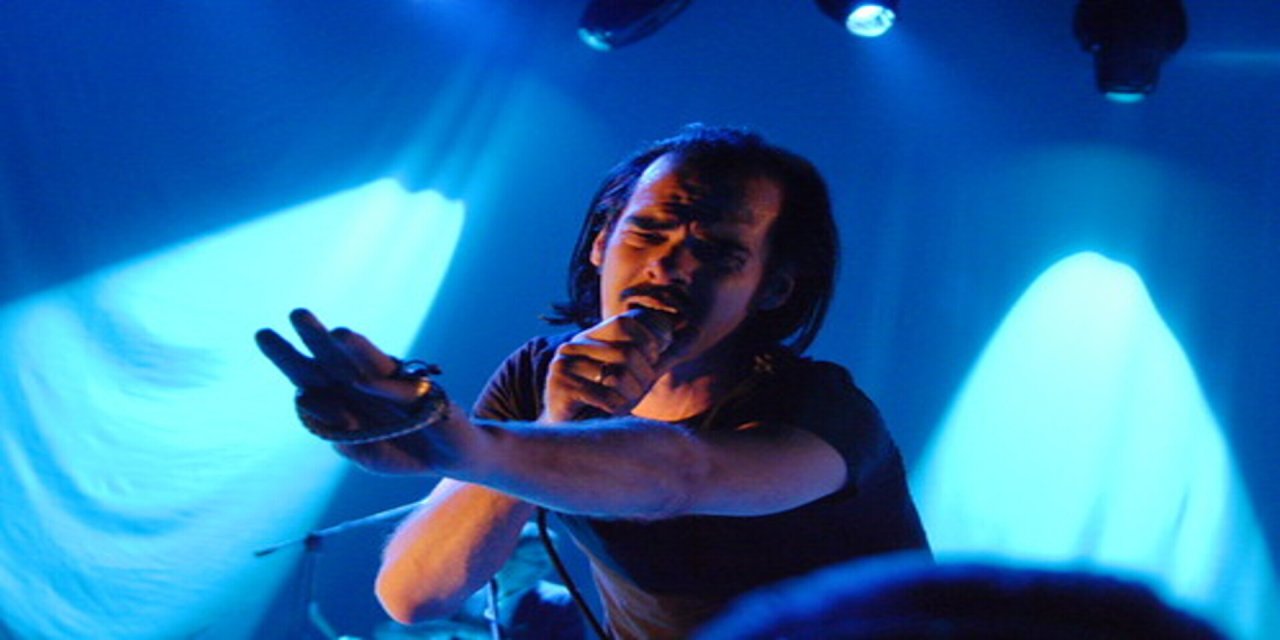 Man with long black hair and mustache singing into microphone with blue backdrop on stage in the background